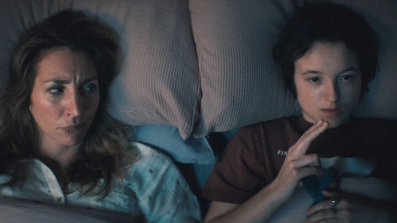 Two people lying side by side in bed on soft, textured pillows, one wearing a patterned pyjama top and the other holding a small blue object, set in a dimly lit, intimate bedroom scene.