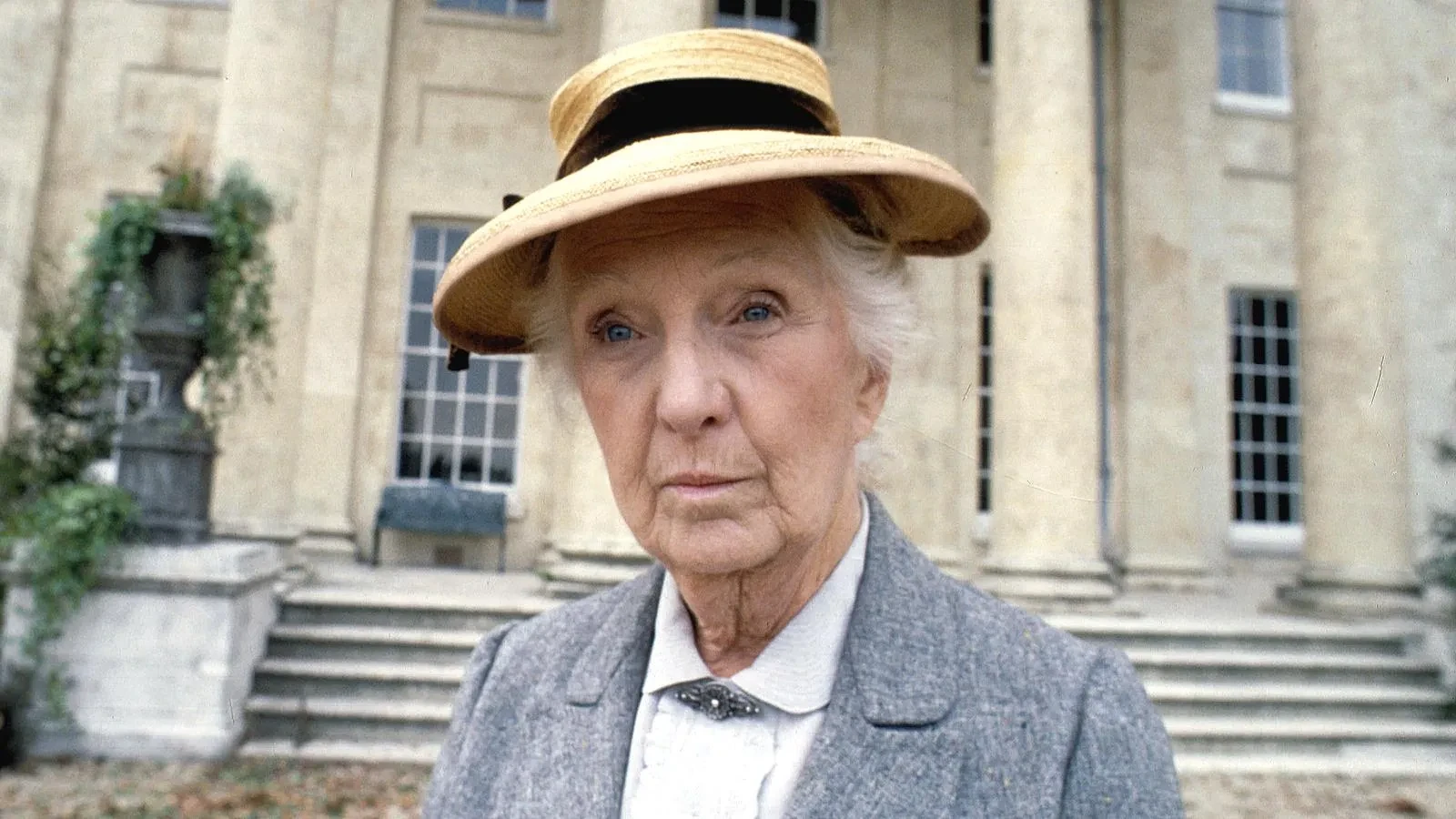 An older woman wearing a straw hat, grey coat and white blouse stands in front of a grand stone manor house with tall columns, large windows and wide steps, creating a classic countryside mystery atmosphere.