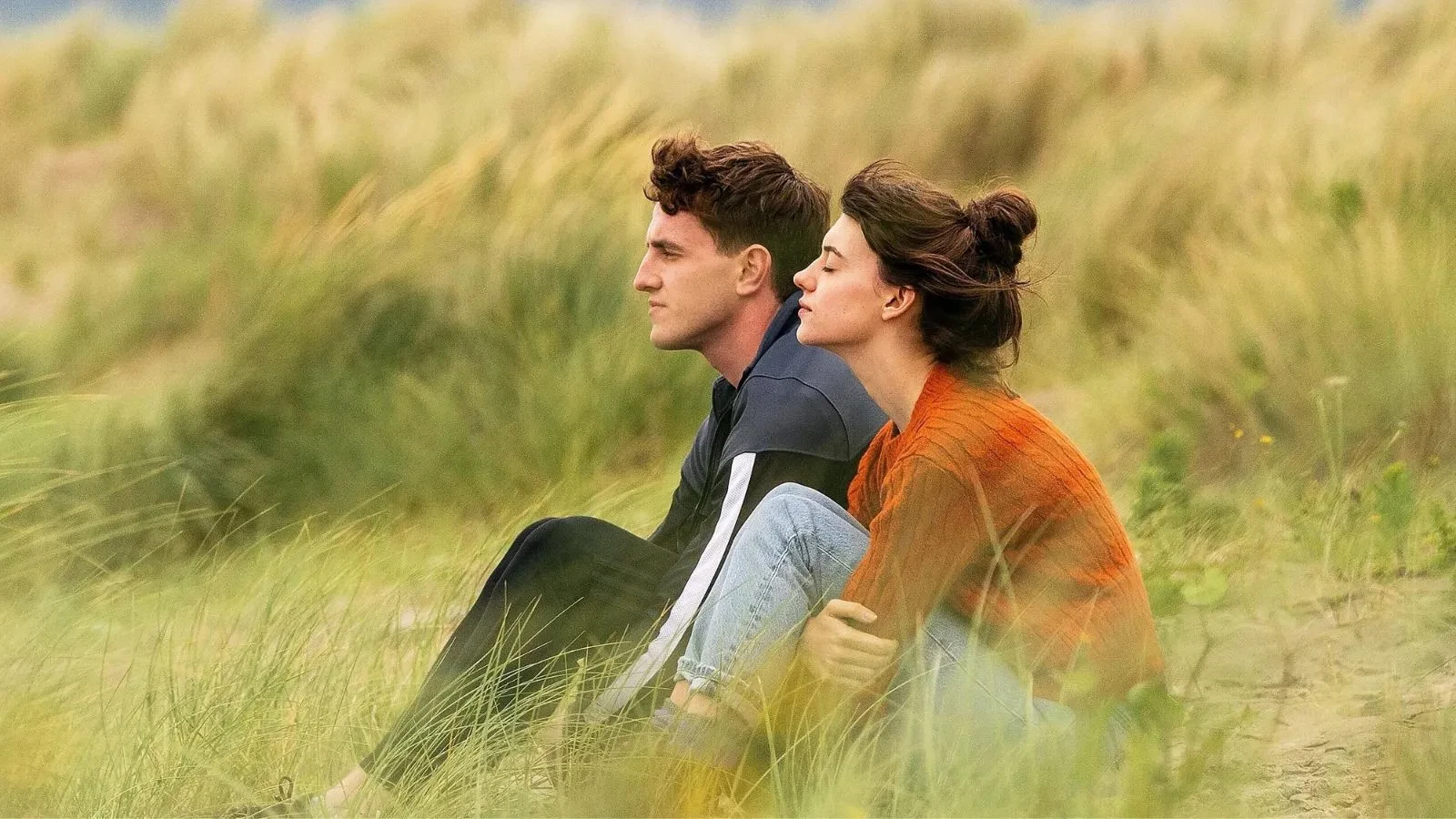 Two people sit close together on grassy sand dunes, dressed in casual clothing. One wears a dark jacket with striped sleeves, and the other wears a burnt orange jumper and light blue jeans. The setting is a natural coastal landscape with tall grass and soft, muted colours, suggesting a calm, outdoor scene.