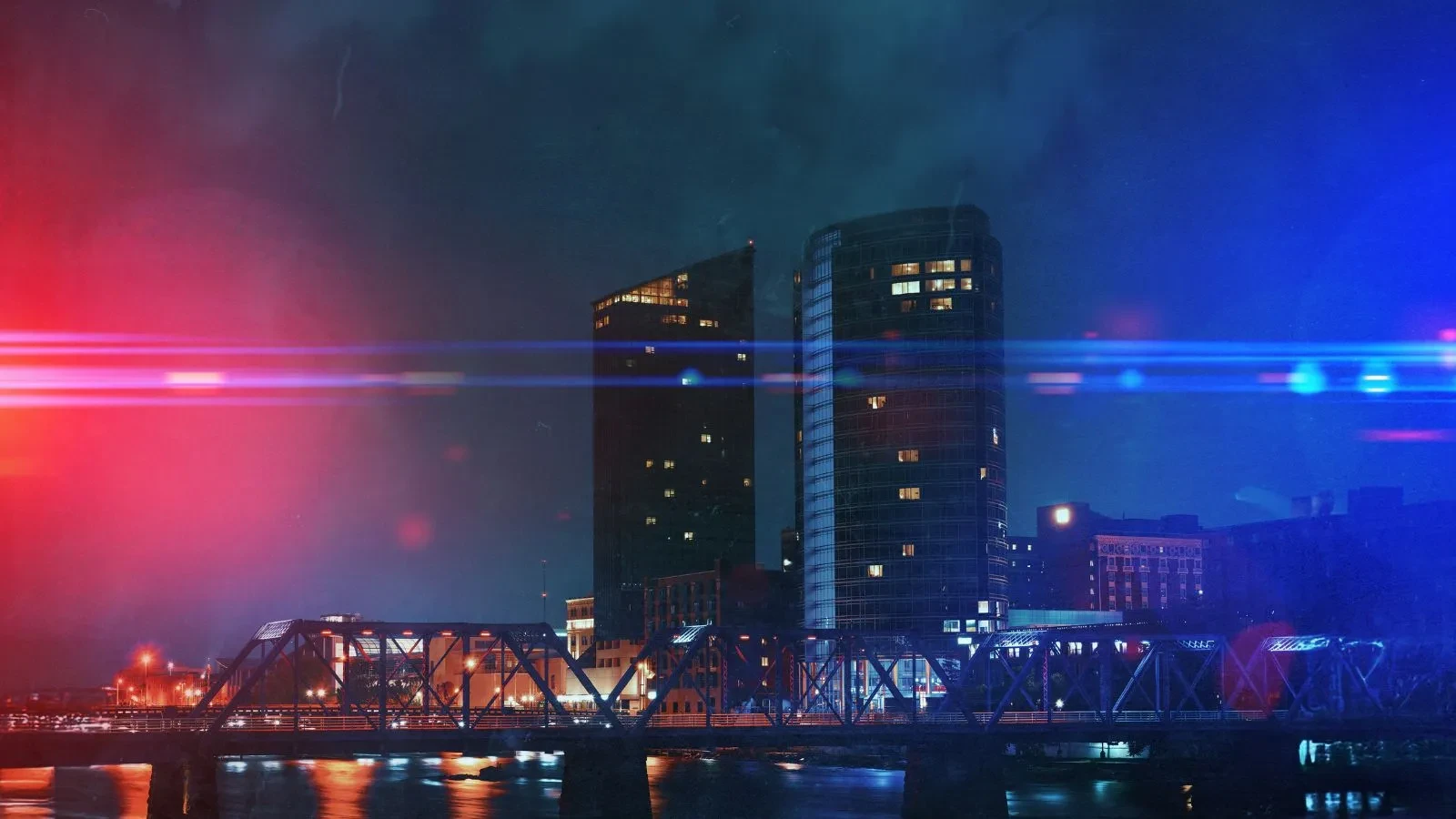 A nighttime cityscape featuring two tall modern buildings and a lit bridge over water. Red and blue light streaks, resembling police lights, frame the image, creating a dramatic effect.
