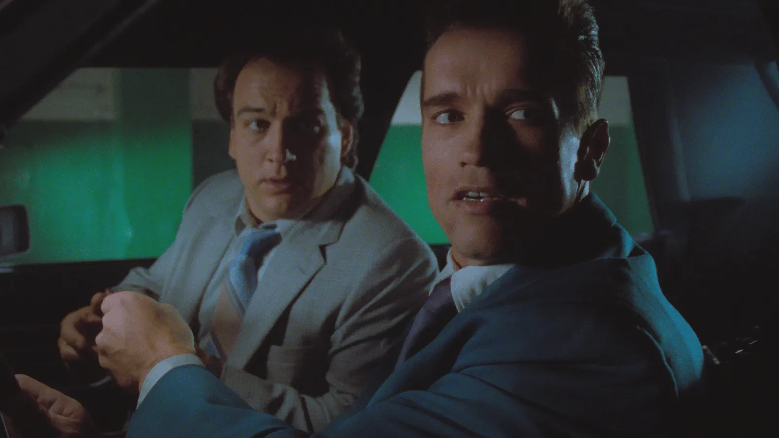 Two people sit inside a car, both wearing suits and ties. One is in the driver’s seat and the other in the passenger seat, and they appear to be talking while the interior is dimly lit with a greenish background visible through the windows.