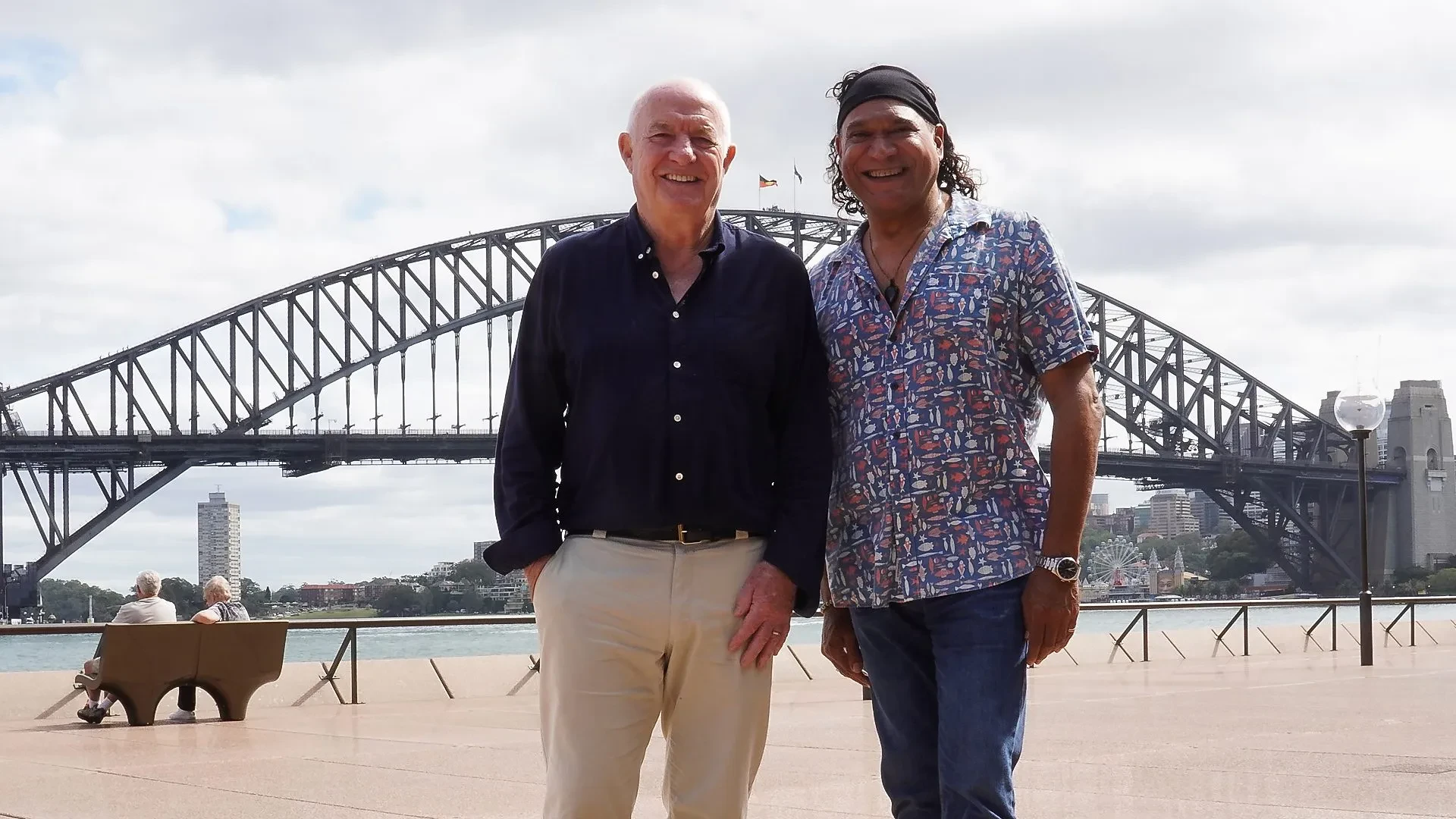 Two people standing on the waterfront in front of Sydney Harbour Bridge on a bright, cloudy day, with the city skyline, water and seated pedestrians visible in the background.