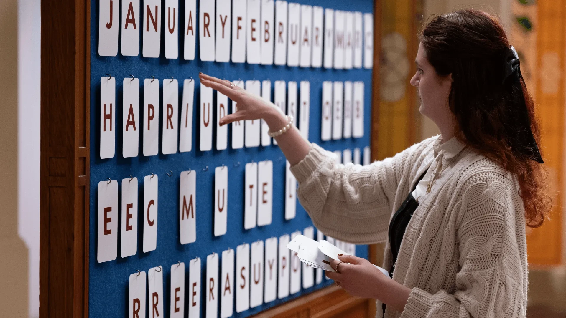 A person wearing a cream knit cardigan arranges letter tiles on a large blue board displaying month names and scrambled words, holding additional tiles in one hand while reaching up to place another. 