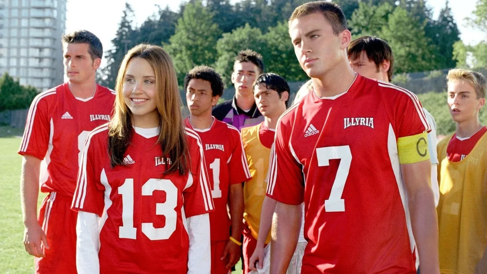 Group of football players wearing matching red Illyria team jerseys with white stripes and numbers, standing together on a grassy sports field with trees and a tall building in the background.