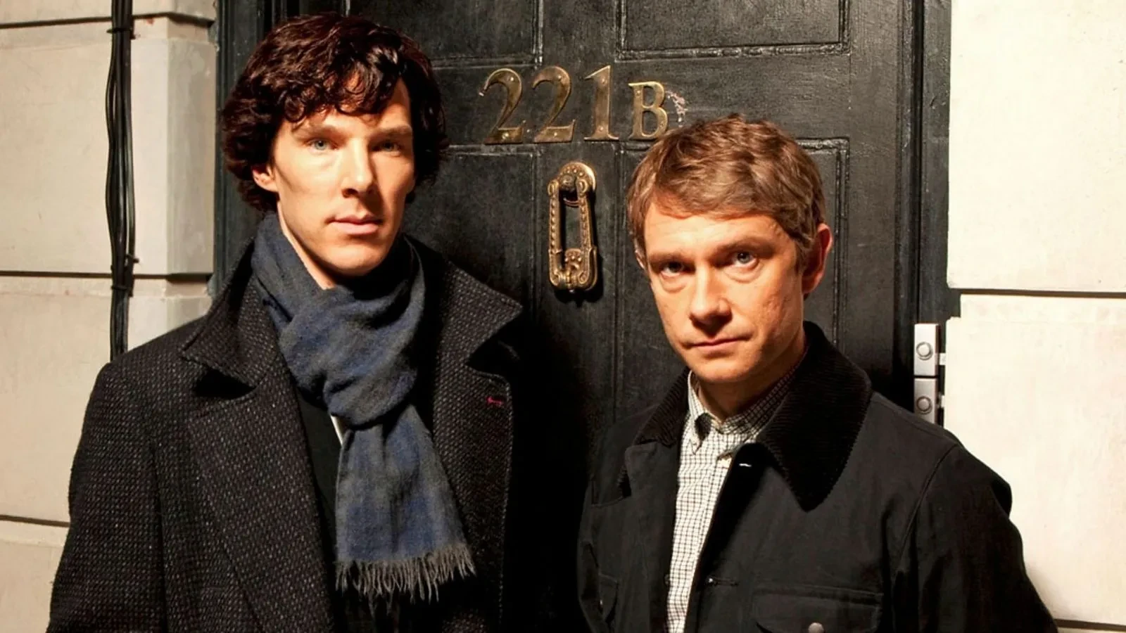 Black door with brass knocker and gold lettering reading ‘221B’, iconic entrance associated with Sherlock Holmes, flanked by two individuals wearing dark coats and standing against a light stone wall.