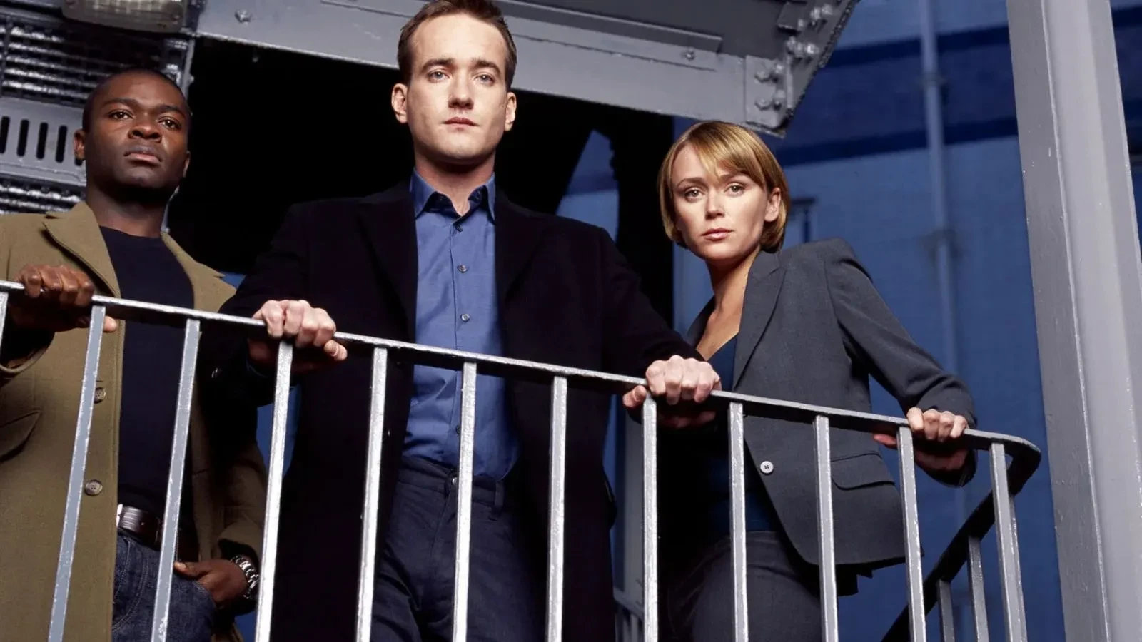 A group of three people in smart business and casual attire standing on an industrial-style metal balcony, looking down with a serious, focused posture against a blue urban backdrop.