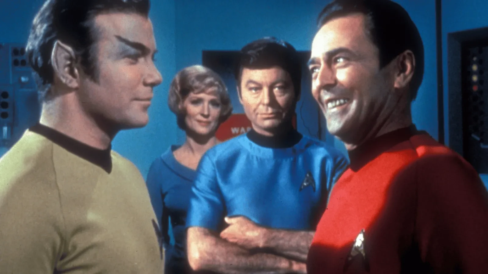 A group of four people in brightly colored scifi uniforms stand together in a futuristic control room. One person wears yellow, one wears red, one wears blue, and another stands slightly behind them in a lighter outfit. Various panels and equipment are visible in the background.
