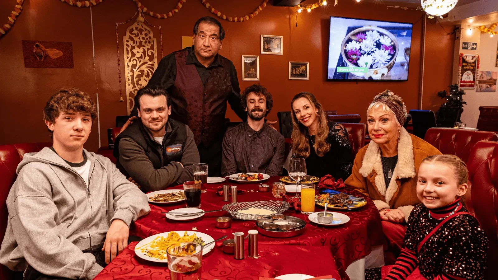 A group of people sit together around a large round table in a warmly lit restaurant, surrounded by red décor and festive string lights. Plates of food, drinks, and a candle sit on the table as they pose for a group photo, with a server standing behind them.  Image 2 Two people stand together in a dimly lit bathroom, brushing their teeth in front of a mirror. Their reflections are visible, and the scene has a warm, intimate, everyday atmosphere.  Image 3 A person wearing a bright green blouse stands in fron