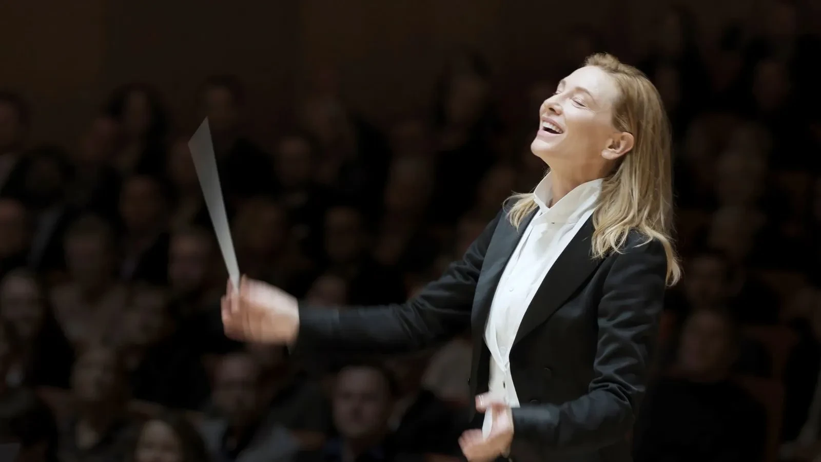 Orchestral conductor wearing a formal black tailcoat and white dress shirt, holding a baton mid-performance against a dark concert hall background.