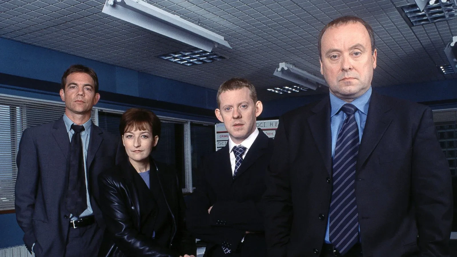 A group of four people dressed in formal business and police-style attire standing inside a blue-lit office with ceiling panels, fluorescent lighting and blinds, creating a serious and professional atmosphere.