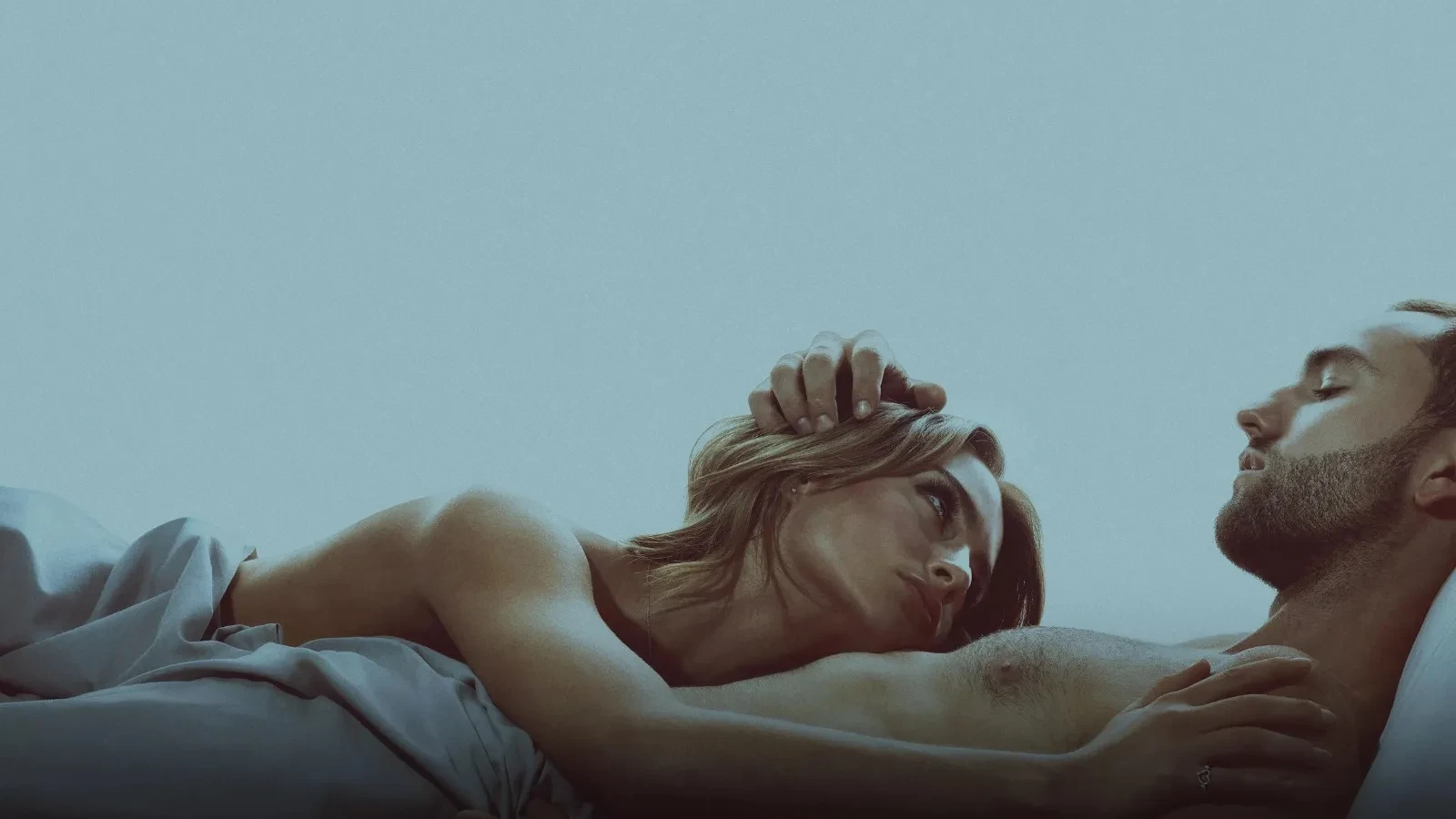 Intimate bedroom scene of a man and woman lying in bed under white sheets, the woman resting her head on the man’s bare chest while gazing thoughtfully at his face, soft natural light creating a calm, emotional and romantic atmosphere.