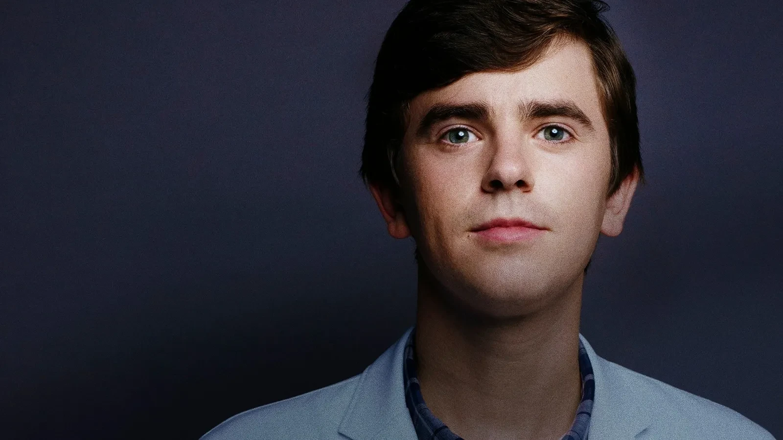 A person wearing a light blue medical-style jacket and a checked shirt standing against a dark, neutral studio backdrop, creating a clean and professional portrait composition.