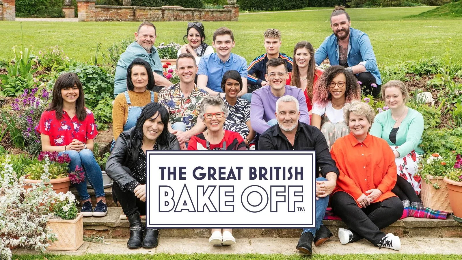 A group of people is sitting and standing outdoors in a garden setting, surrounded by flowers and greenery. They are dressed in colorful, casual clothing. In front of them is a large sign that reads “The Great British Bake Off.”