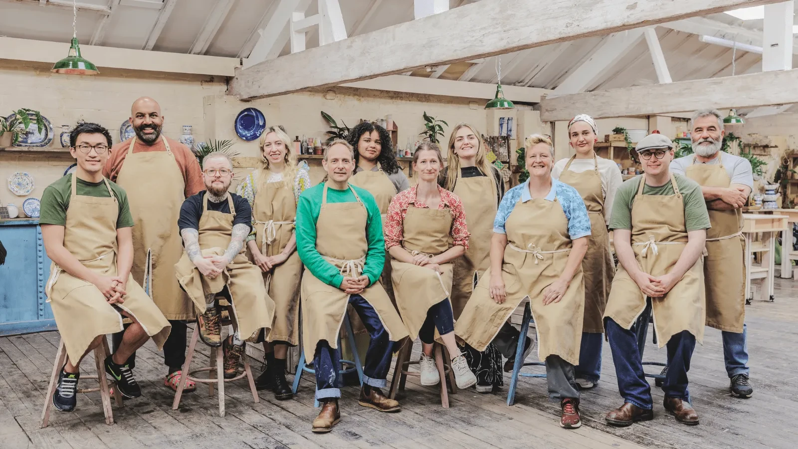 A group of people wearing aprons sit and stand together in a rustic workshop filled with pottery, plants, and wooden beams overhead.