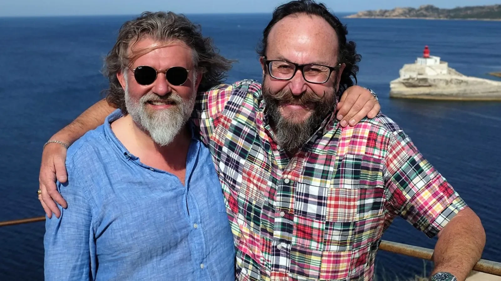 Two people standing arm‑in‑arm on a sunny coastal viewpoint overlooking deep blue Mediterranean waters, with a small white lighthouse perched on a rocky outcrop in the background.