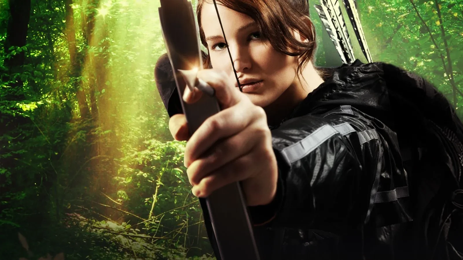 Close-up of a person in a black jacket drawing a bow and arrow in a sunlit forest.