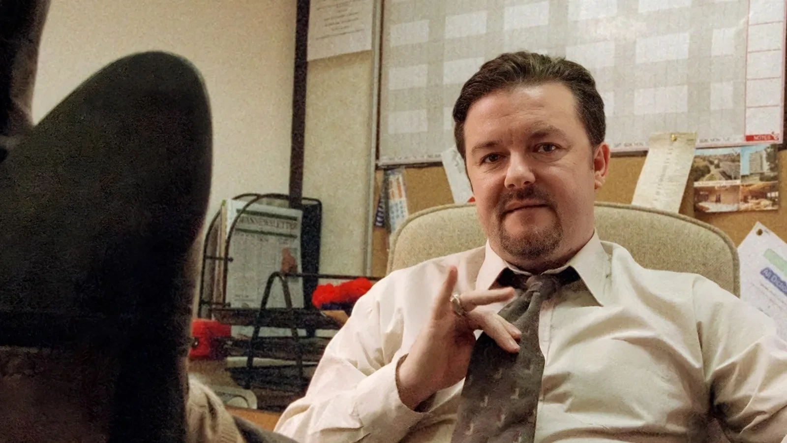 A man in an office setting sits back in his chair with his feet up on the desk, adjusting his tie in a relaxed yet authoritative pose, surrounded by paperwork, noticeboards and filing trays.