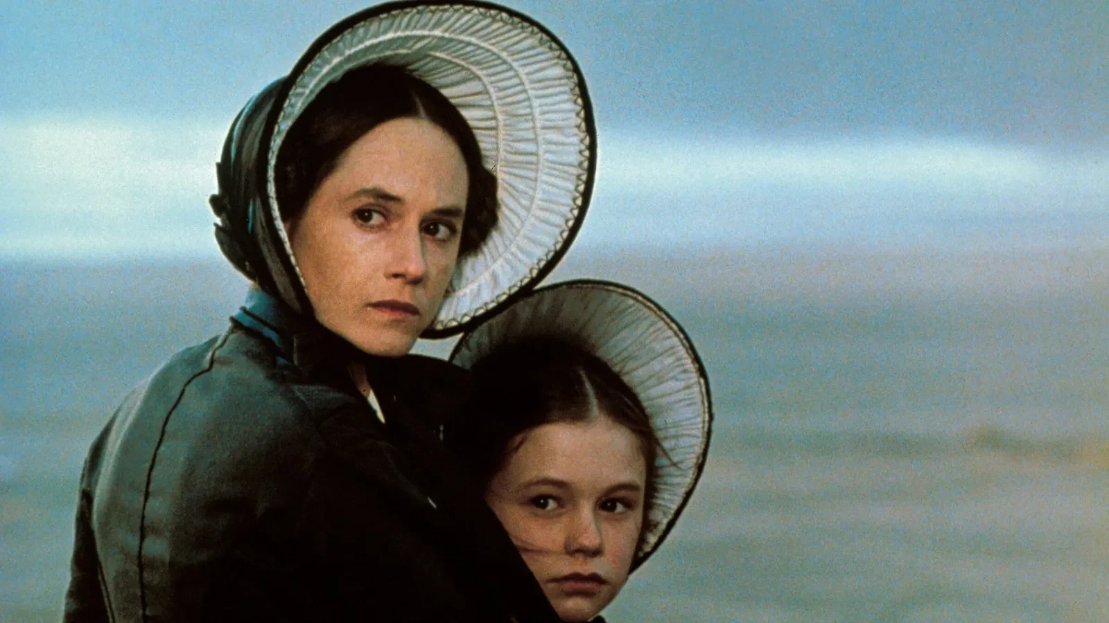 Two figures wearing traditional 19th‑century bonnets and dark period clothing stand closely together by a windswept coastline, framed against a soft, muted seascape under an overcast sky.