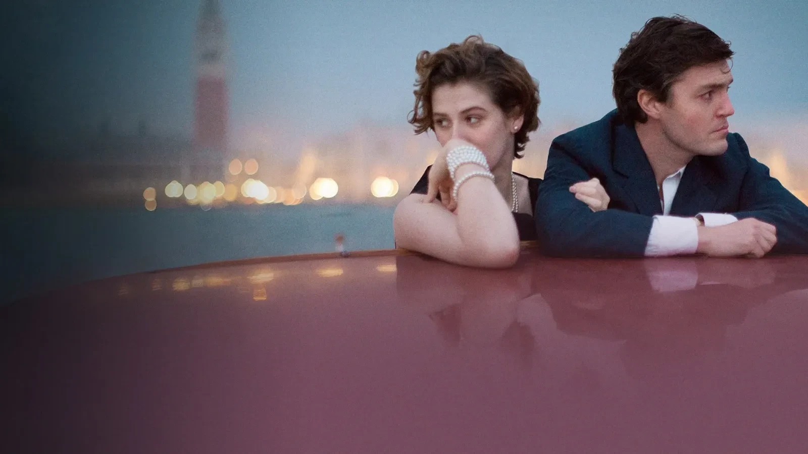 A man and a woman leaning on the roof of a boat at dusk, with soft city lights and a tall tower in the distant, hazy background, creating a calm and atmospheric waterfront scene.