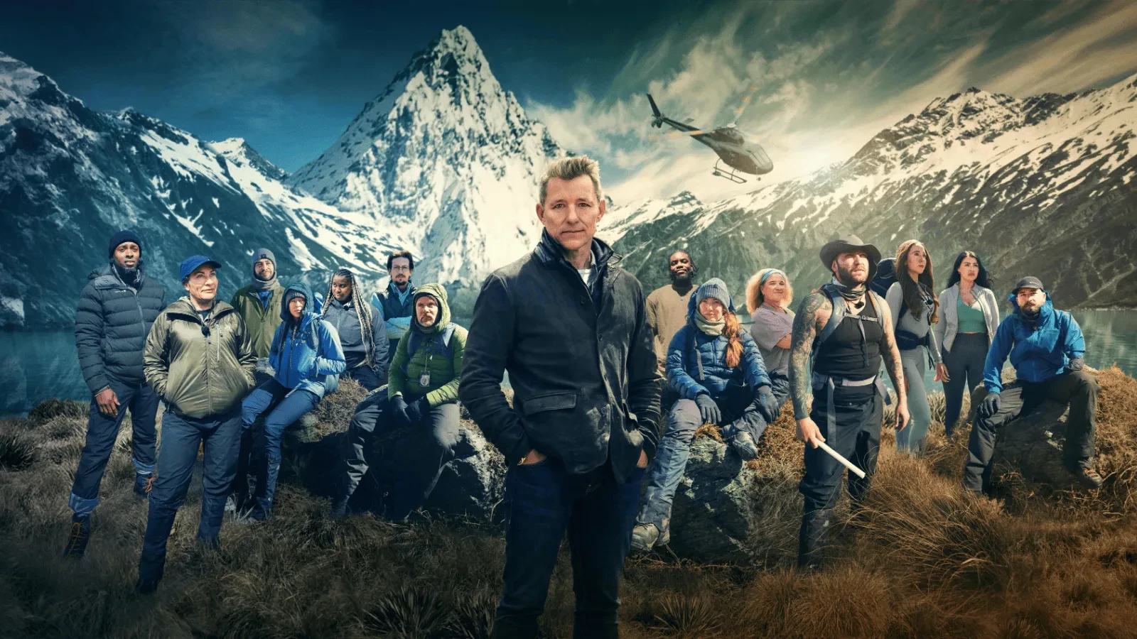 A large group of adventurers stands in rugged outdoor gear against dramatic snow‑capped mountains with a helicopter in the sky.