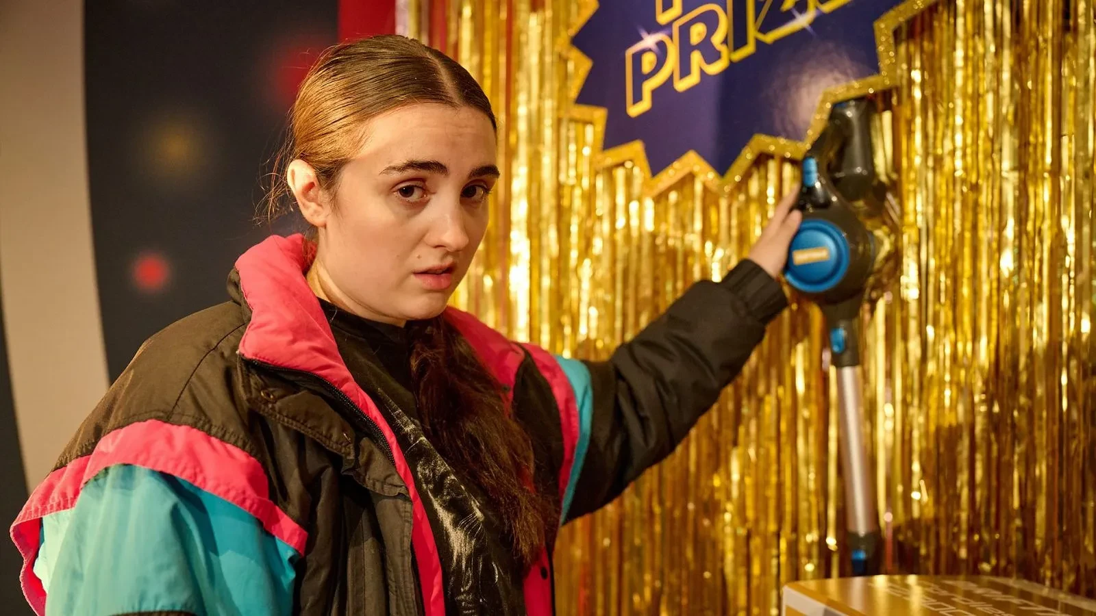 Person wearing a retro-style jacket with teal and pink panels stands in front of a gold tinsel backdrop, holding a cordless vacuum cleaner mounted on a display. A sign above reads “PRIZE” in bold letters, suggesting a game show or prize giveaway setting.