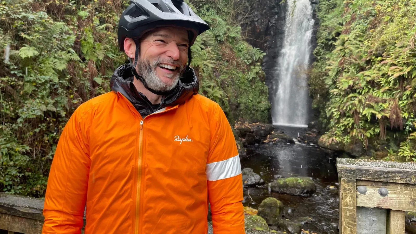 A cyclist wearing a bright orange Rapha jacket and a black helmet stands on a wooden viewing platform in front of a tall forest waterfall, surrounded by lush green foliage and moss‑covered rocks.