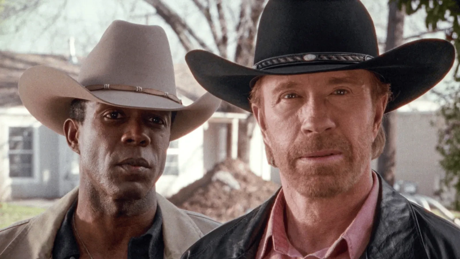 Two people wearing cowboy hats stand outdoors in front of a house and trees. One is dressed in a light-coloured hat and jacket, and the other wears a black hat with a leather jacket over a collared shirt