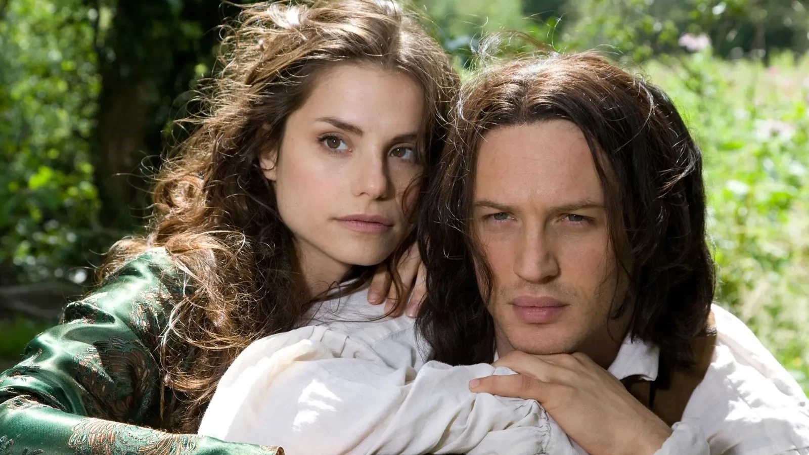 A close-up outdoor scene showing two people embracing tenderly, dressed in period-style clothing with flowing hair, set against a lush green countryside backdrop suggestive of a romantic and dramatic atmosphere.