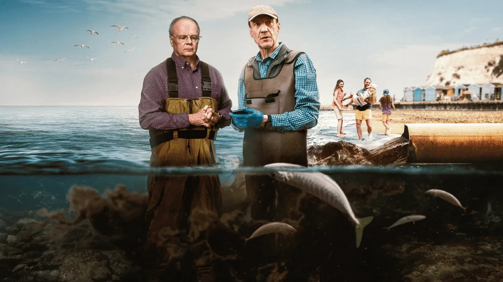 Two individuals wearing waders stand near the shoreline discussing marine conditions, with fish swimming underwater in the foreground and a coastal scene with beach huts, cliffs, and people near the water in the background.