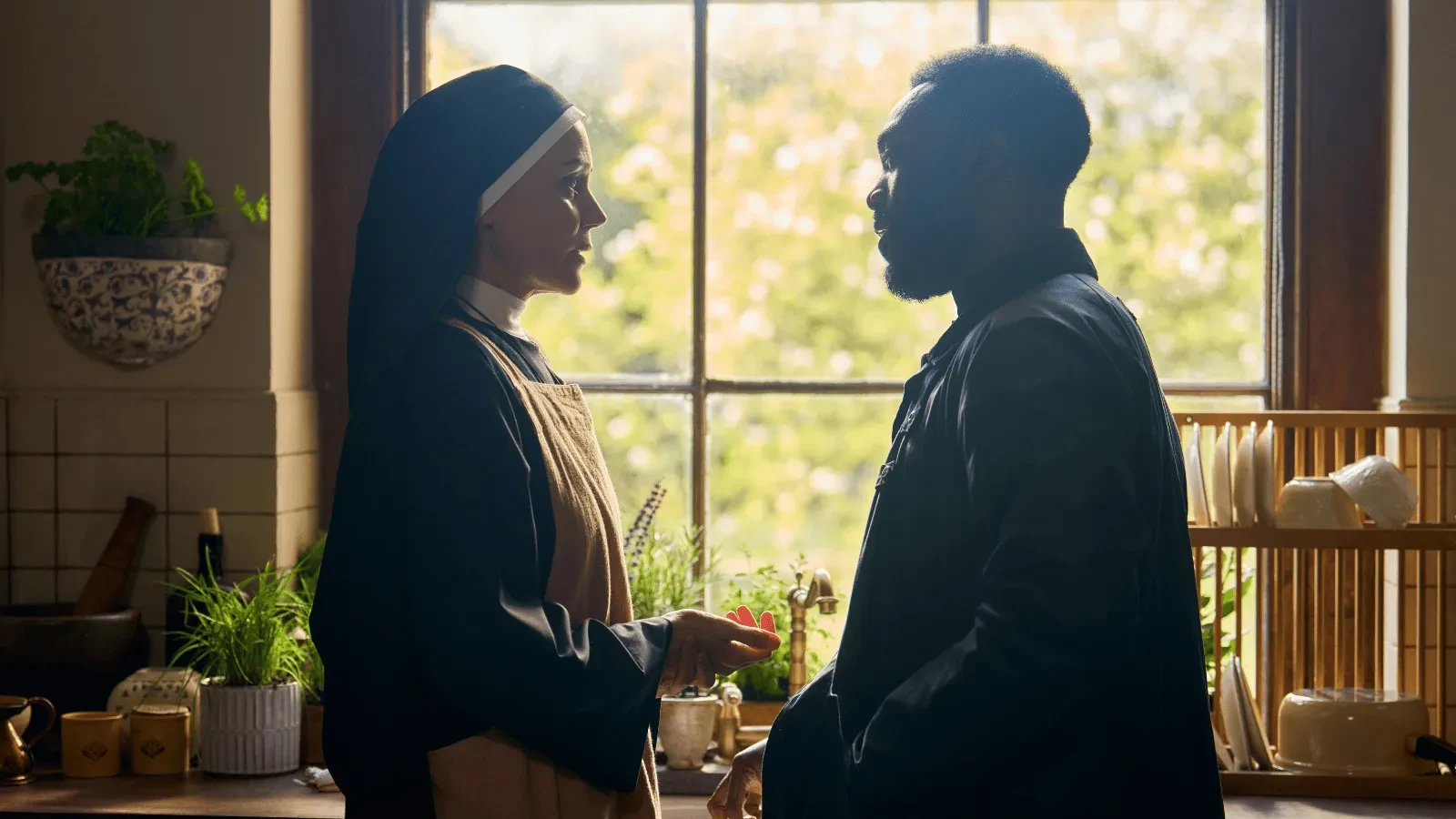 Two people standing in a sunlit kitchen by a large window, having a conversation surrounded by plants and cooking utensils