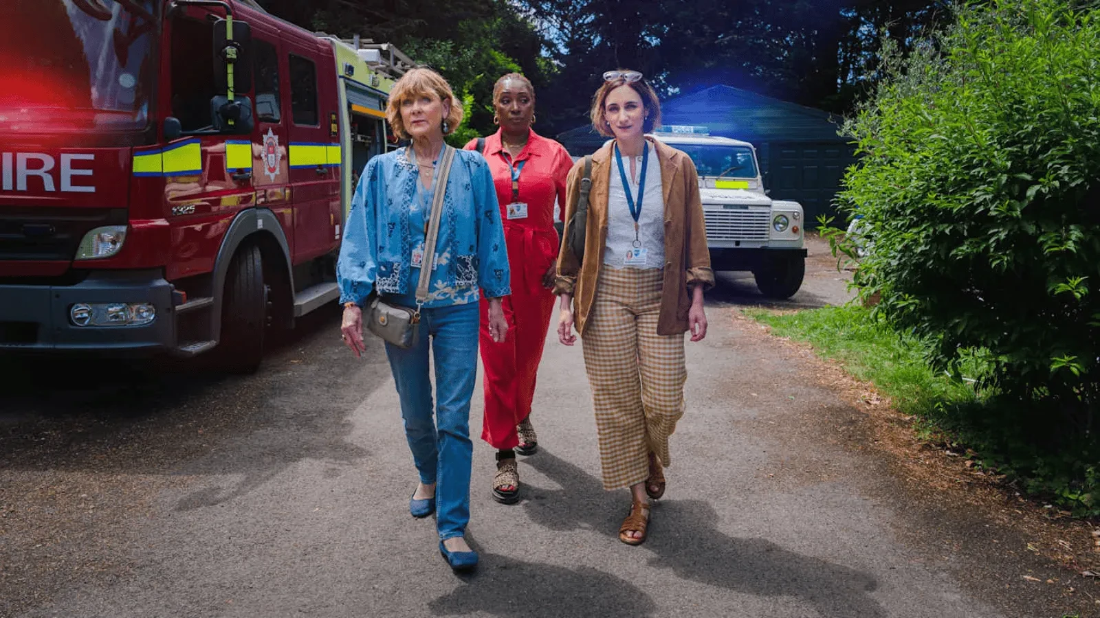 Three individuals walking along a road past a fire engine and an emergency vehicle, wearing lanyards and outdoor clothing in a suburban setting