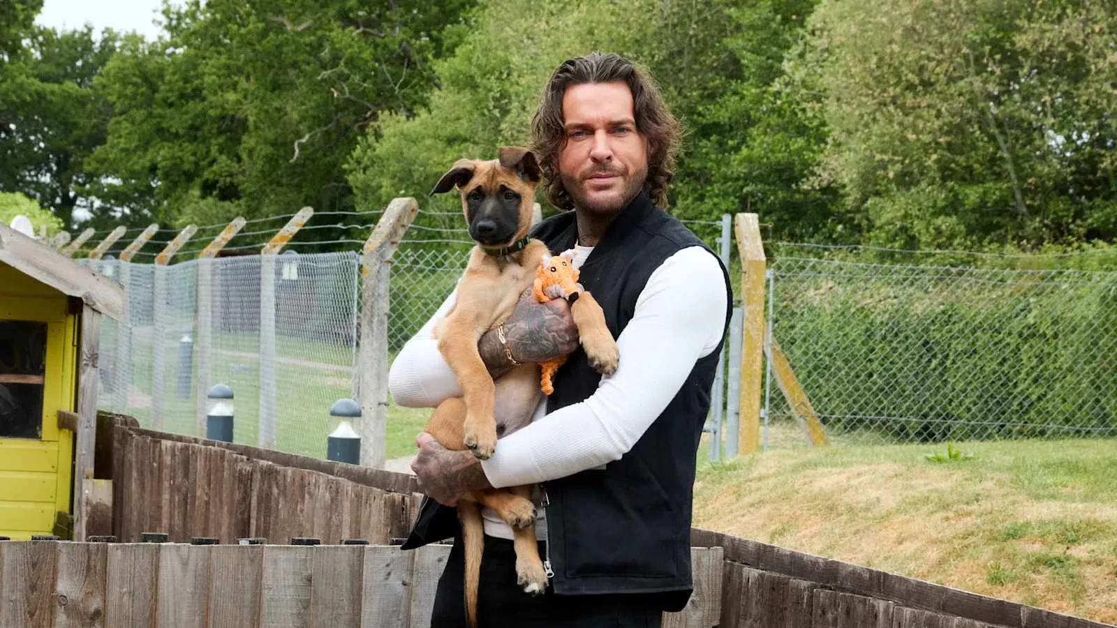 A person stands outdoors in a fenced grassy area, holding a young brown dog in their arms. The dog has its front paws resting on the person’s arm and is clutching a small orange toy. Trees, a wooden fence, and a wire-mesh perimeter fence create a peaceful, shelter-like setting in the background