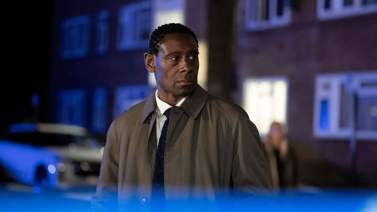 Person wearing a trench coat standing outside a residential building at night, with police lights casting a blue glow