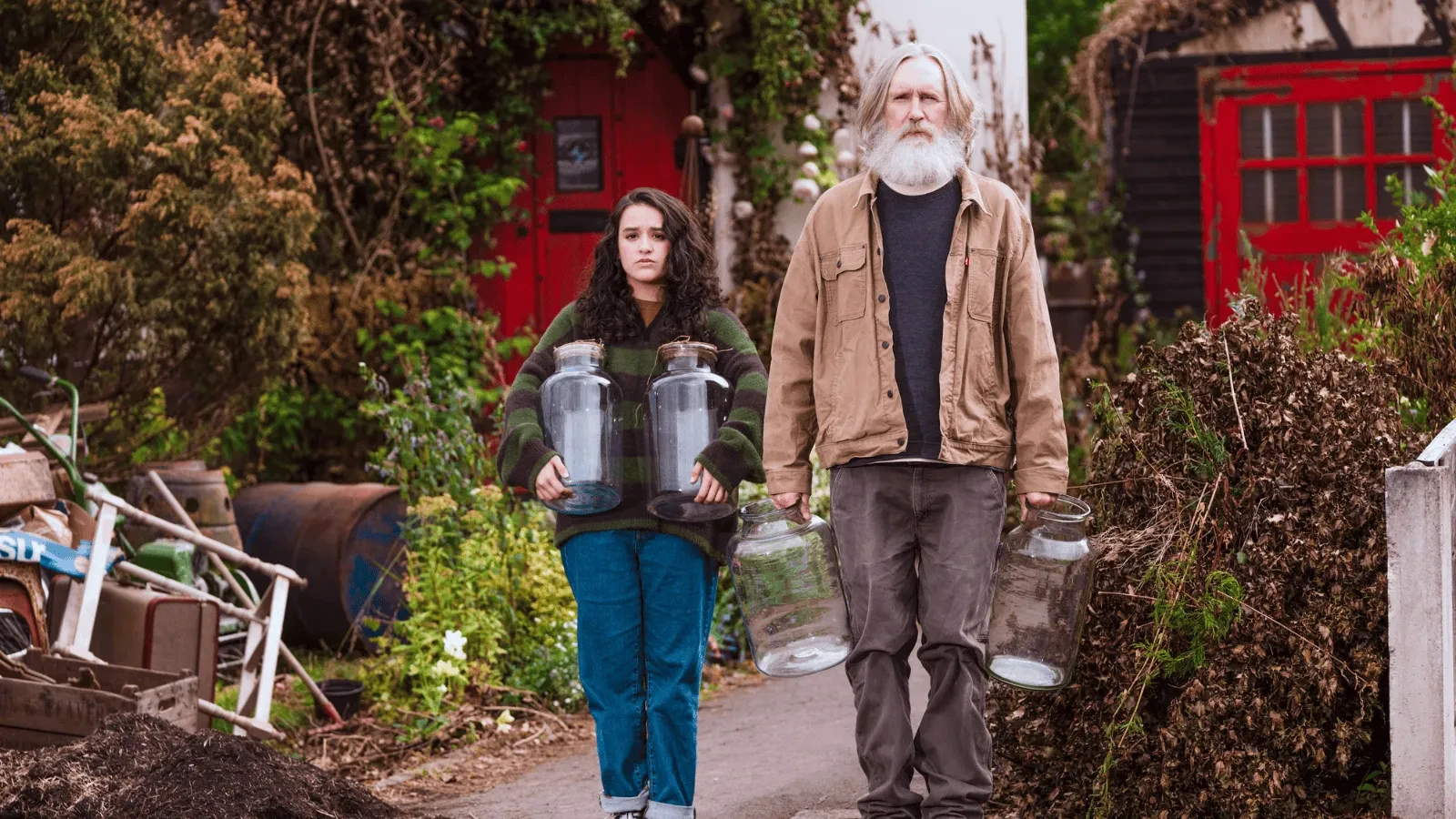 Two people walking down a rustic path carrying oversized glass jars, surrounded by overgrown plants, old buildings, and scattered outdoor items.