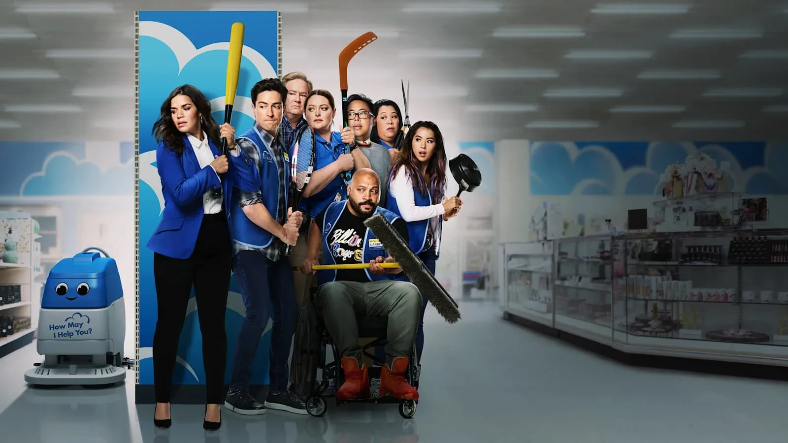 A group of retail employees pose together in a brightly lit megastore aisle, each holding humorous makeshift “weapons” like a bat, hockey stick, broom, and mop. They wear matching blue store uniforms, standing in front of colourful display panels. A small smiling floor‑cleaning robot sits to the side, and shelves stocked with products line the background, creating a playful and chaotic workplace scene