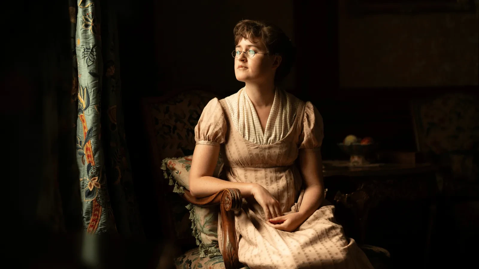 Woman in a light, vintage-style dress sitting on an antique chair near a softly lit window with patterned curtains
