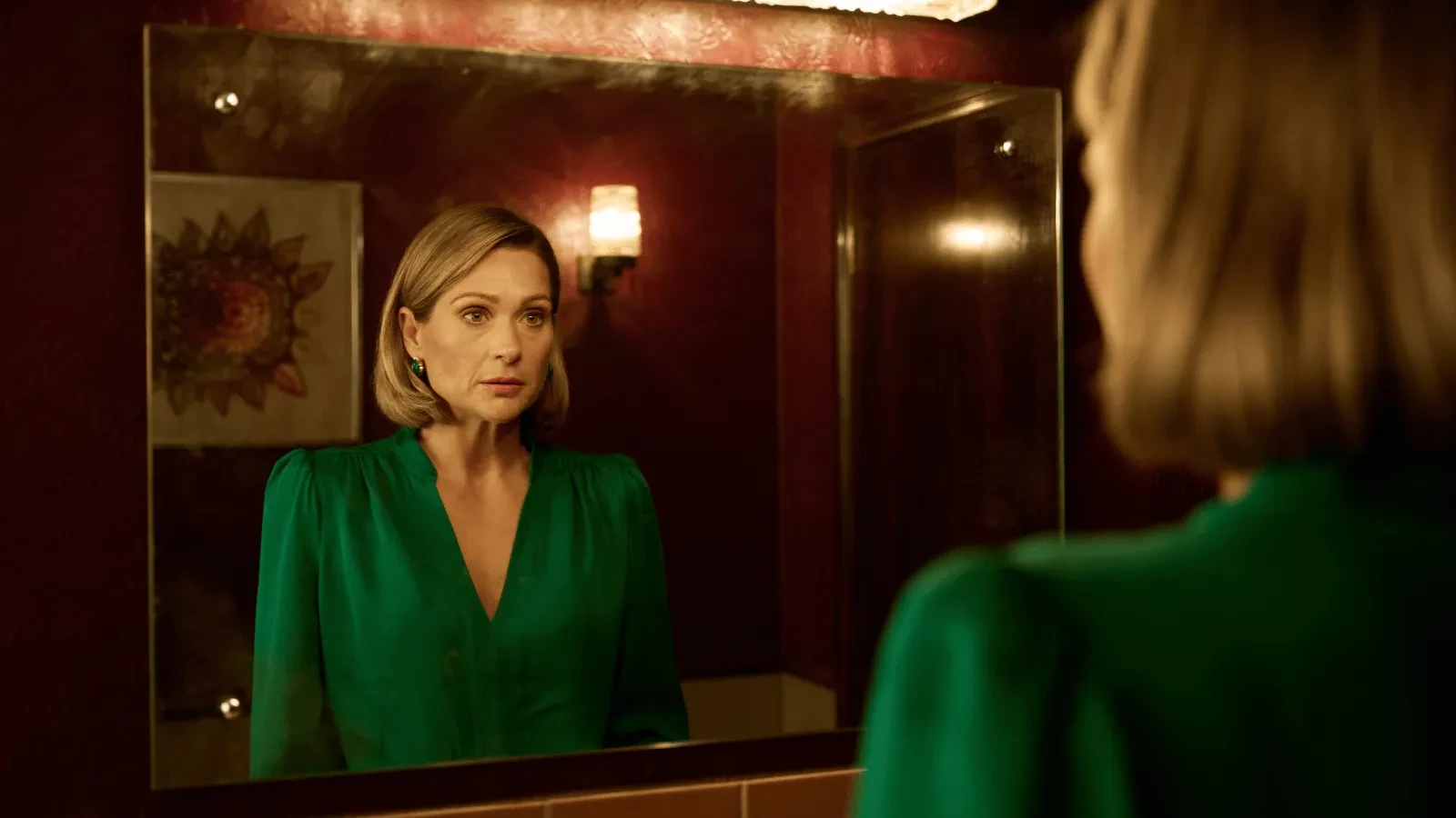 Woman in a green dress standing in a warmly lit bathroom, looking at her reflection in the mirror.