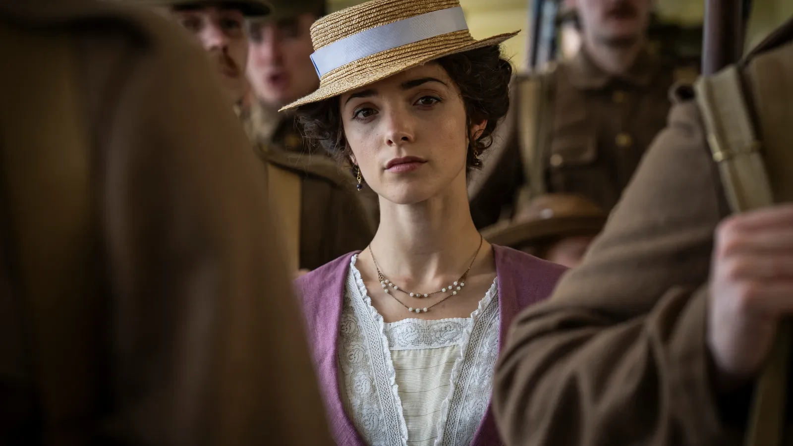 A person dressed in period-style clothing, including a straw hat with a ribbon and a layered lace-trimmed outfit, stands among uniformed soldiers in a crowded indoor setting. 