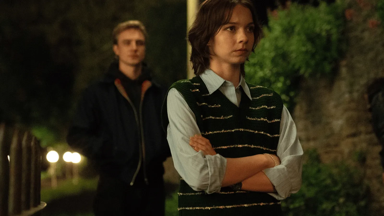Two people standing outdoors at night on a dimly lit path, with one person in a striped sweater vest crossing their arms while another stands slightly behind near a garden wall