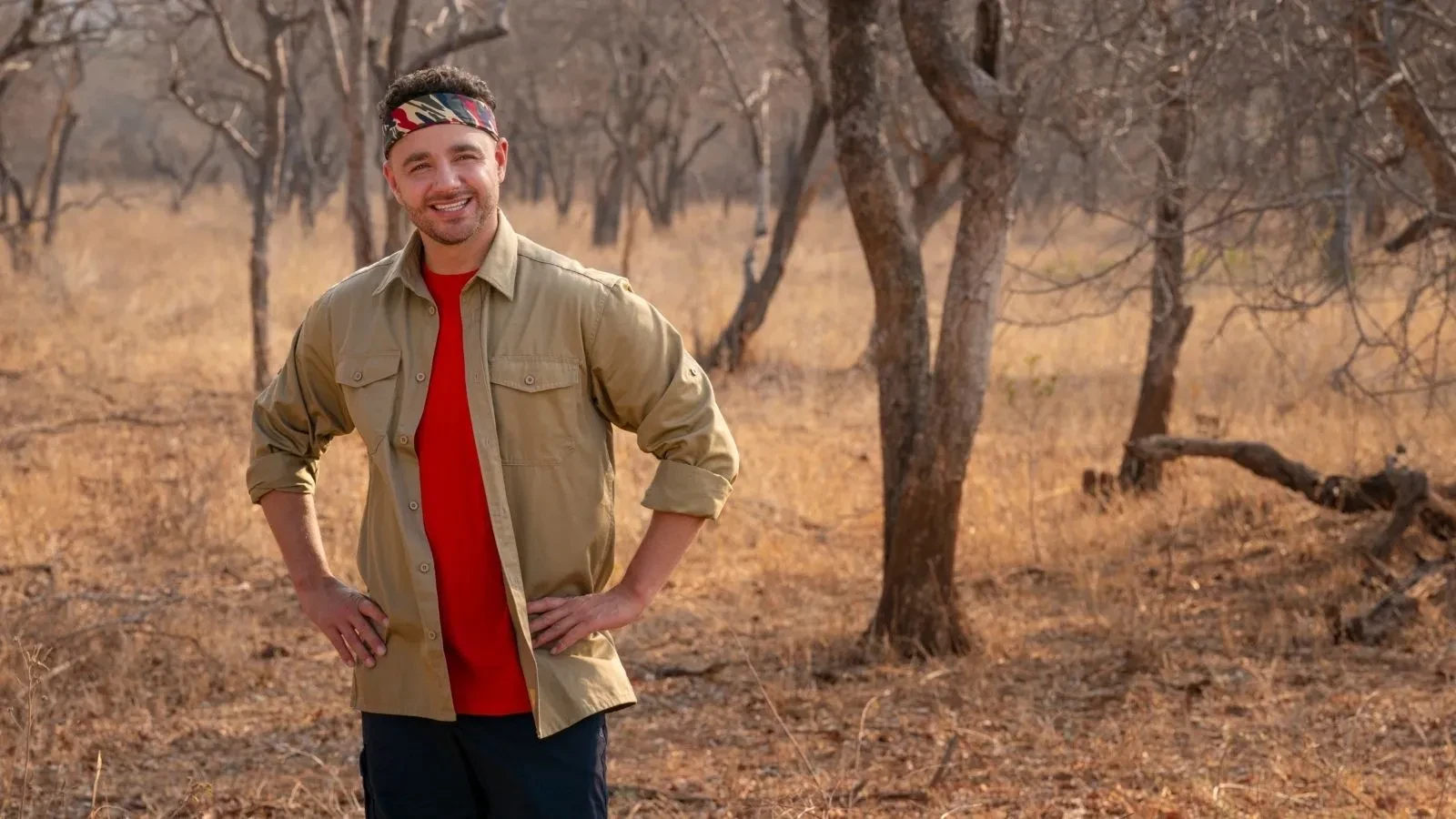 Adam Thomas standing in a dry South African savannah landscape wearing a red T‑shirt and khaki shirt with hands on hips.