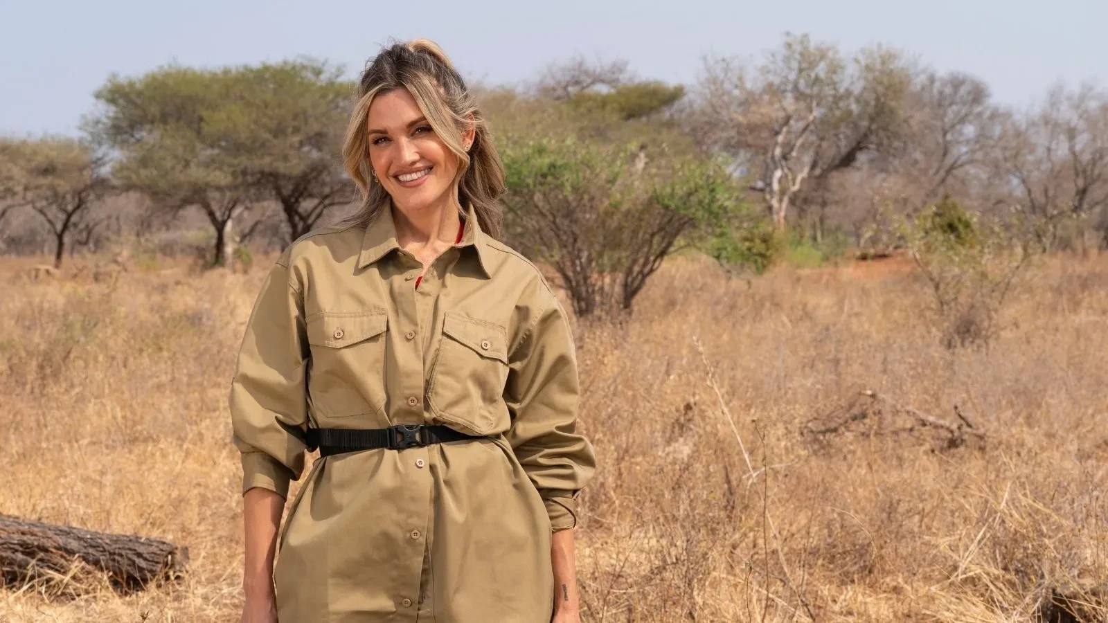 Ashley Roberts standing in the South African wilderness wearing a belted khaki shirt and red top, with bushes and trees behind her.