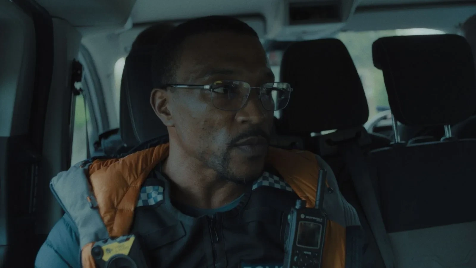 Individual wearing a high‑visibility police vest seated inside a vehicle with communication equipment visible.