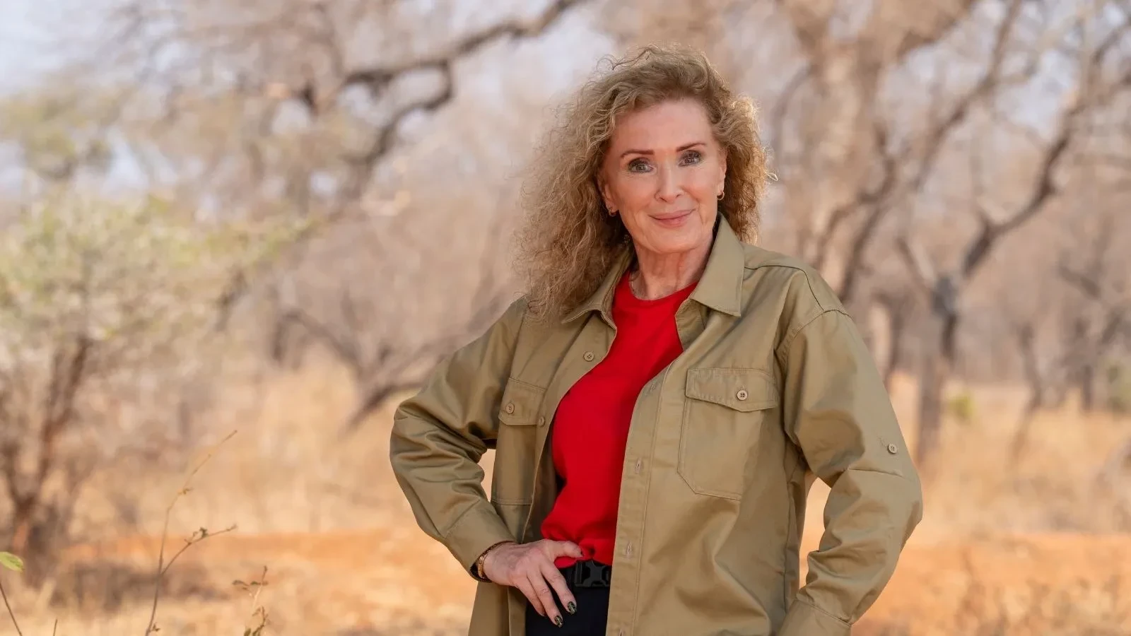 Beverly Callard standing in a dry woodland setting in South Africa, wearing a red top and khaki shirt with one hand on her hip.