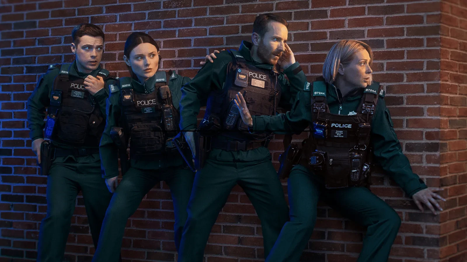 Four uniformed police officers positioned tactically against a brick wall, appearing to prepare for action. 