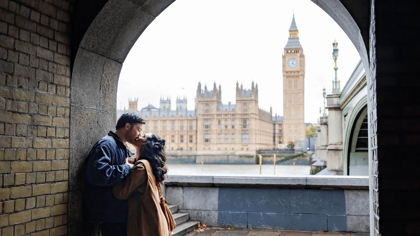 Two people standing closely together under a brick archway beside the River Thames, looking out toward the Palace of Westminster and Big Ben in London.