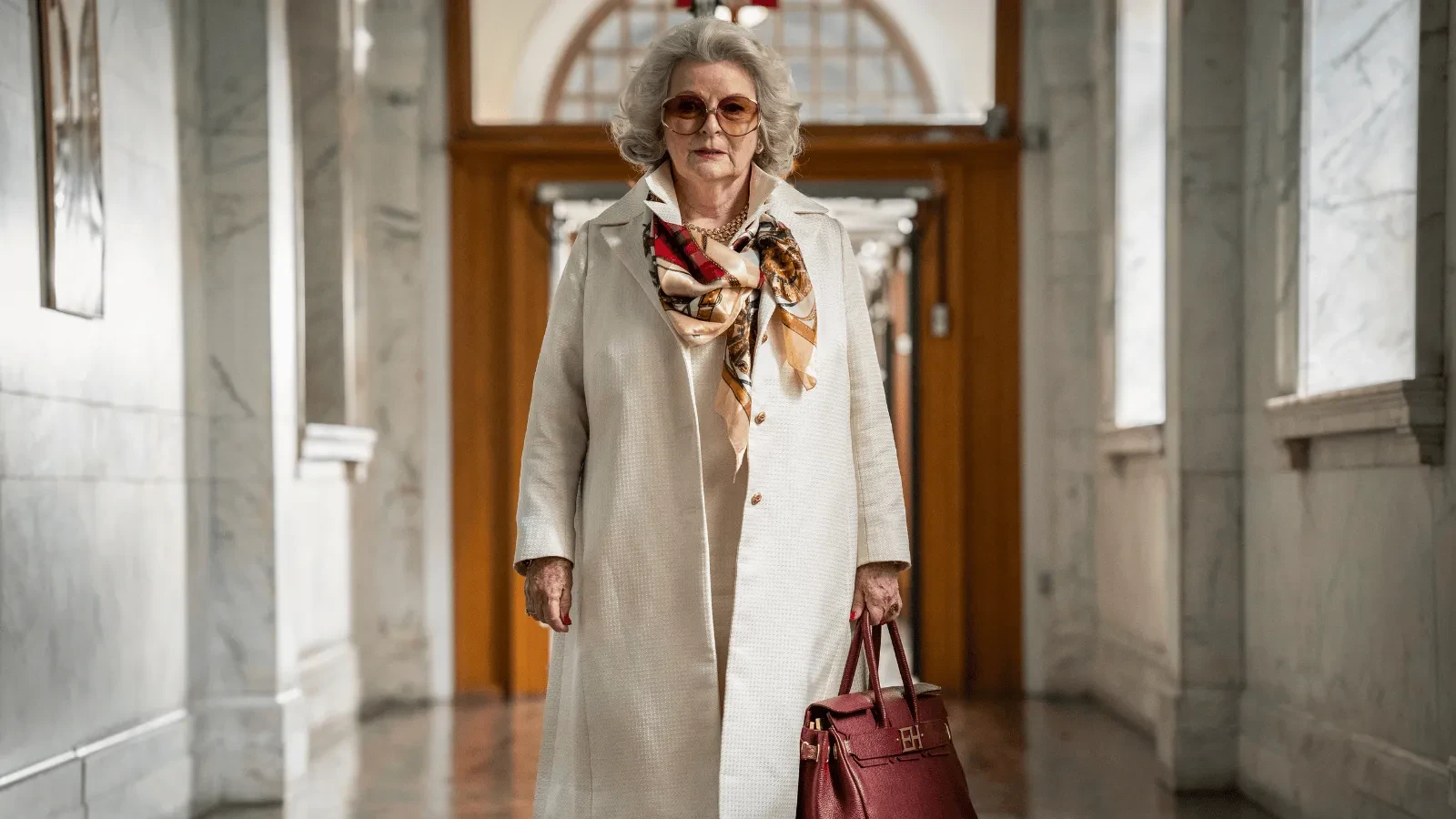 An older woman wearing a long cream coat and patterned silk scarf walks through a marble courthouse hallway, carrying a large red handbag 