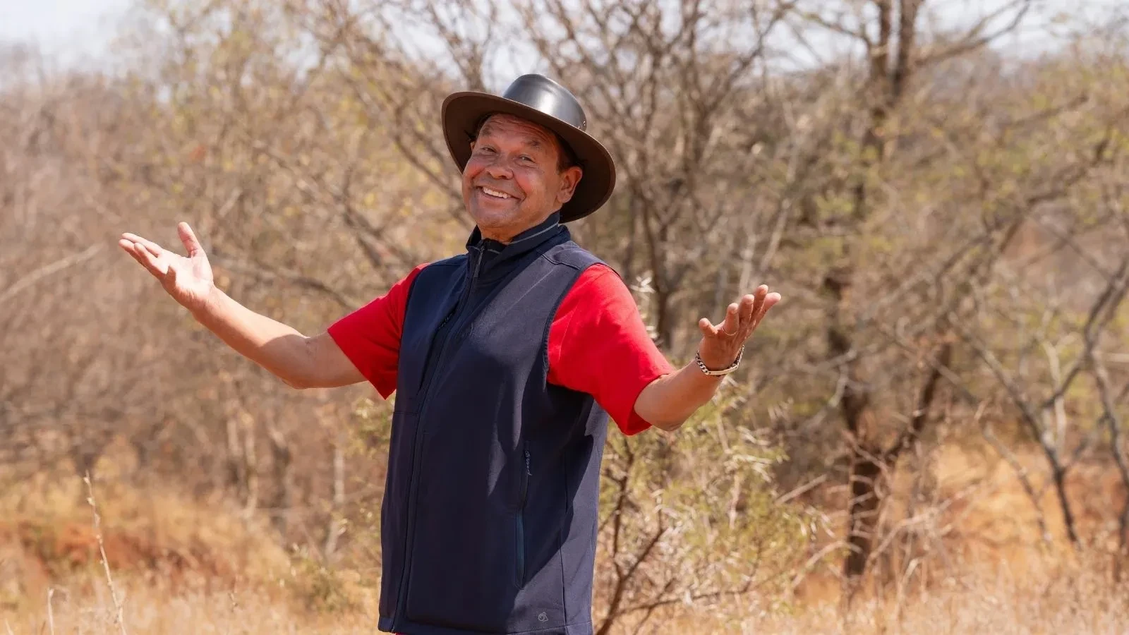 Craig Charles standing with folded arms in a wooded South African setting, wearing a khaki shirt and navy vest.