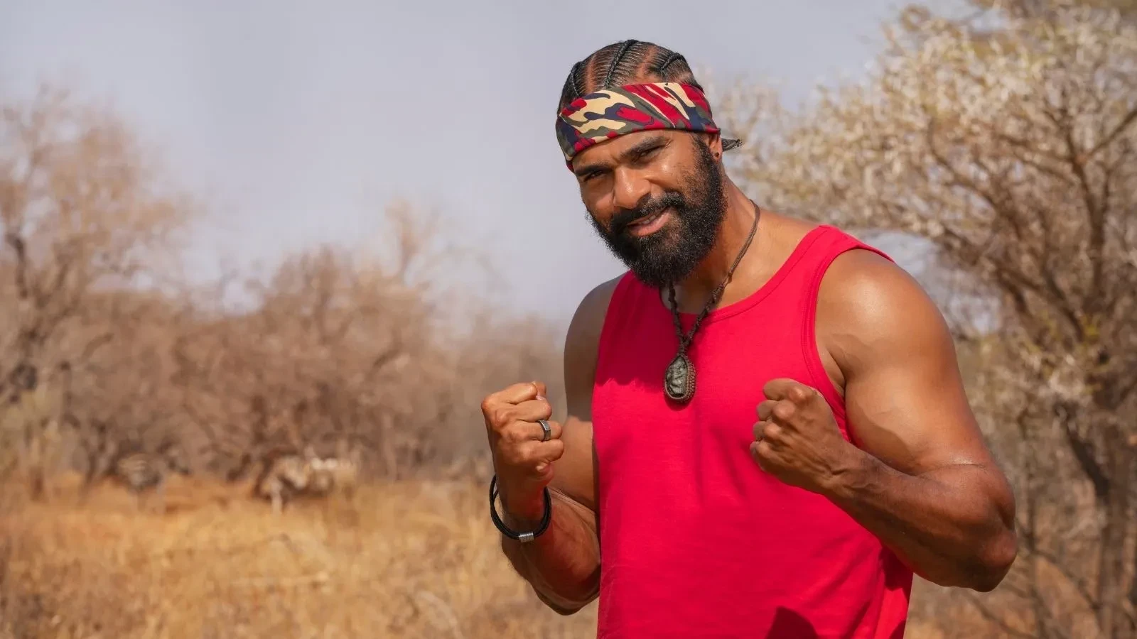 David Haye standing in a dry savannah area wearing a red sleeveless top and bandana, holding his fists up in a powerful stance.