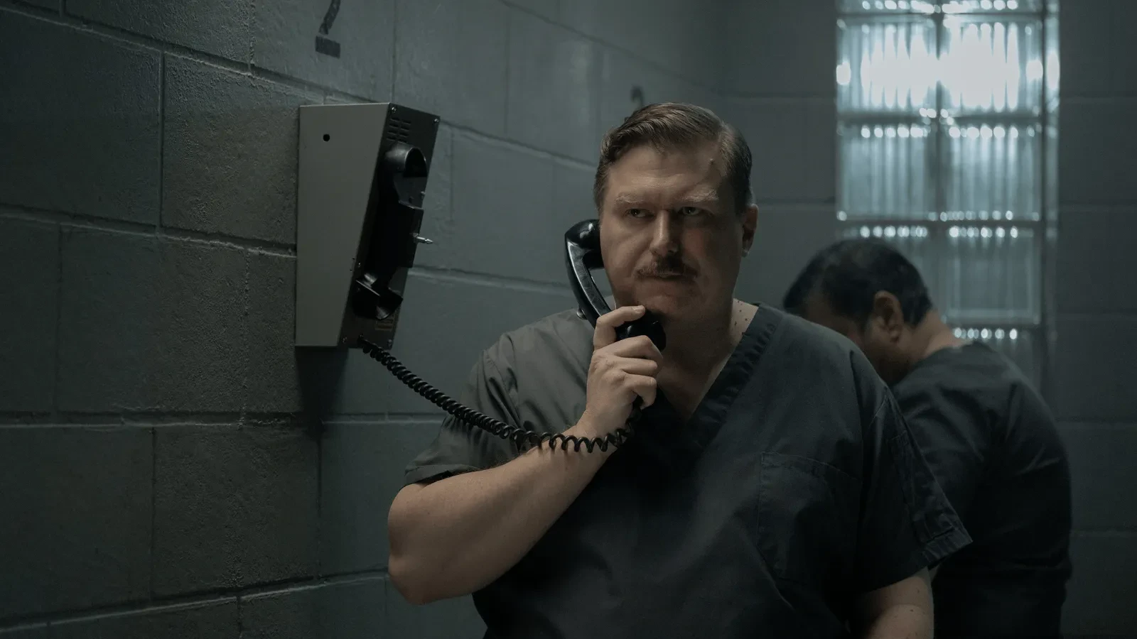 A person wearing dark prison scrubs stands in a cell-like room holding a wall-mounted telephone, while another person stands in the background near a concrete wall and glass-block window. 