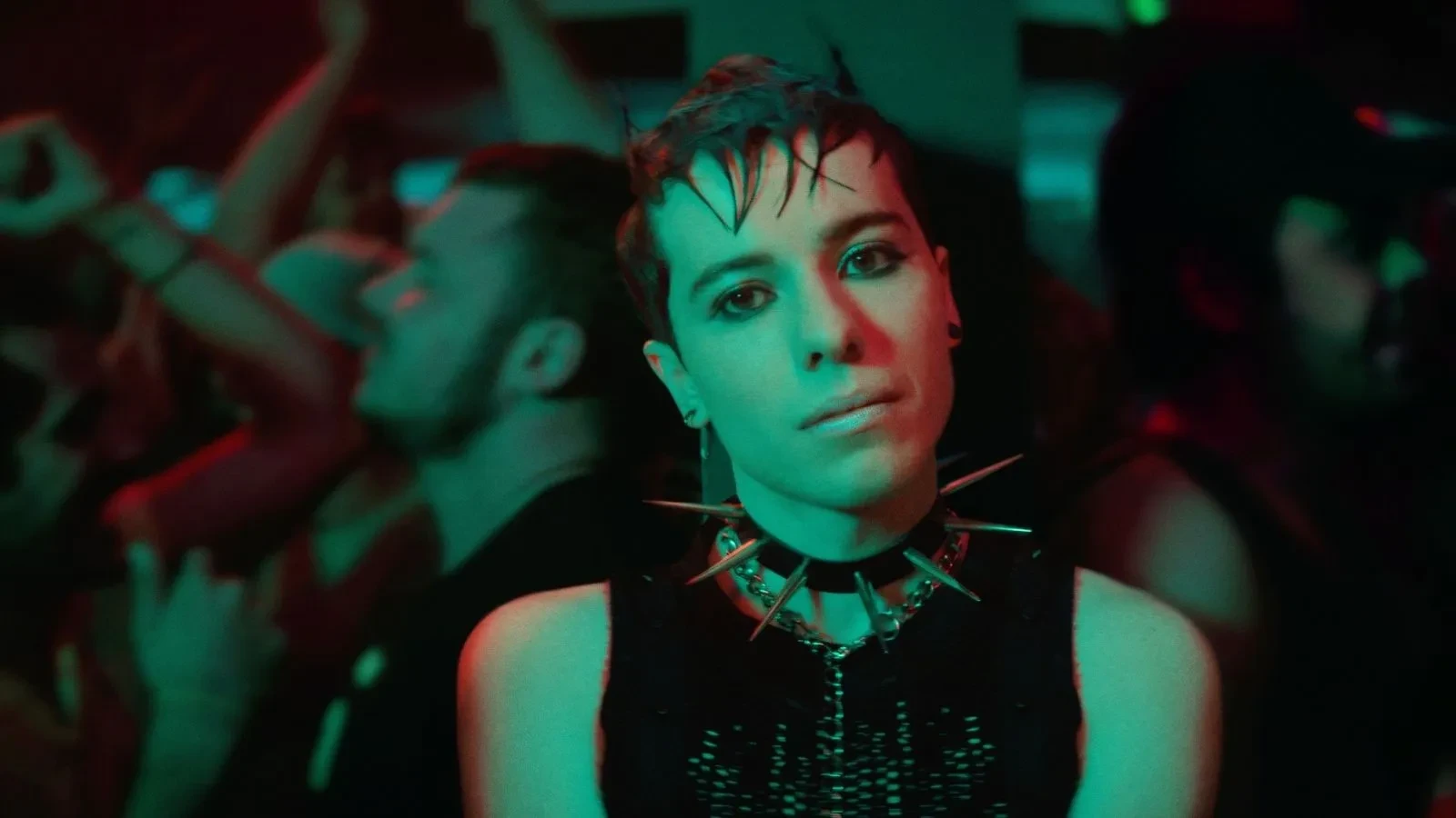 Person in punk-style clothing with a spiked choker standing in a crowded nightclub with neon lighting.